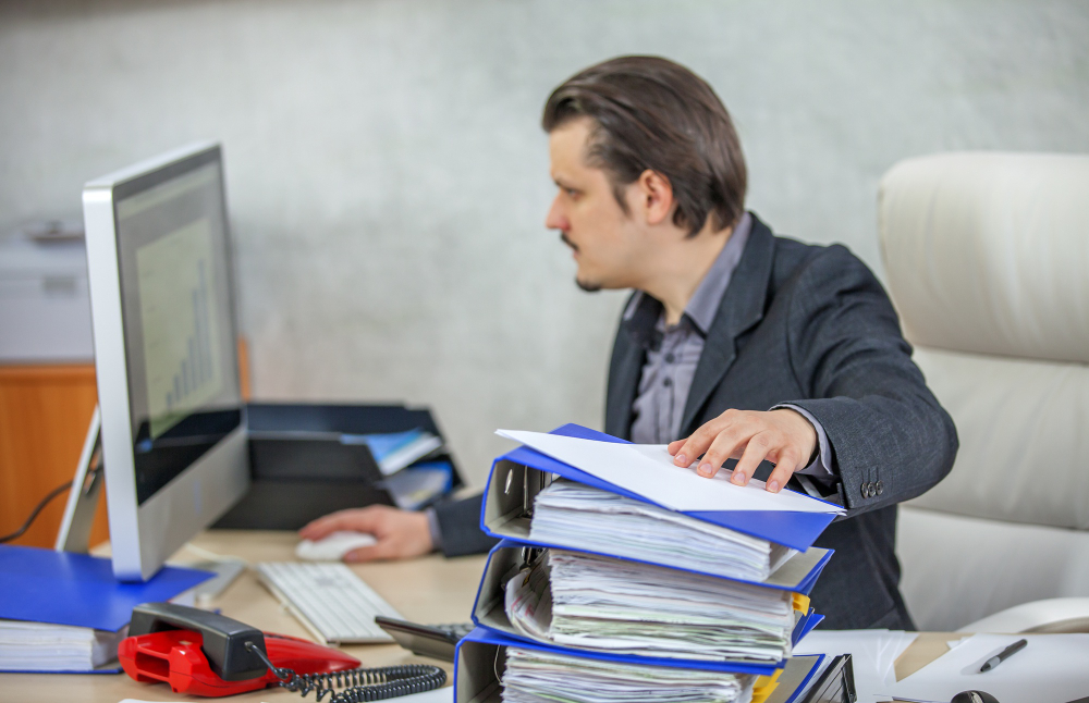 A man in a formal suit is sitting at a desk, focused on a computer screen and thinking “Why is it important to report incidents in the workplace”. The office workspace has various office supplies, including a phone, files, and a computer monitor displaying a graph.