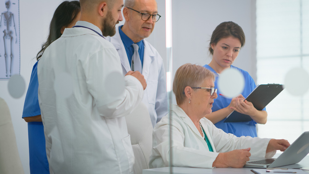 A group of healthcare professionals, including doctors and nurses, gather in a hospital meeting room. One female doctor is seated at a desk, typing on a laptop, while another nurse takes notes on a clipboard. The team appears to be discussing the incident reporting procedure.