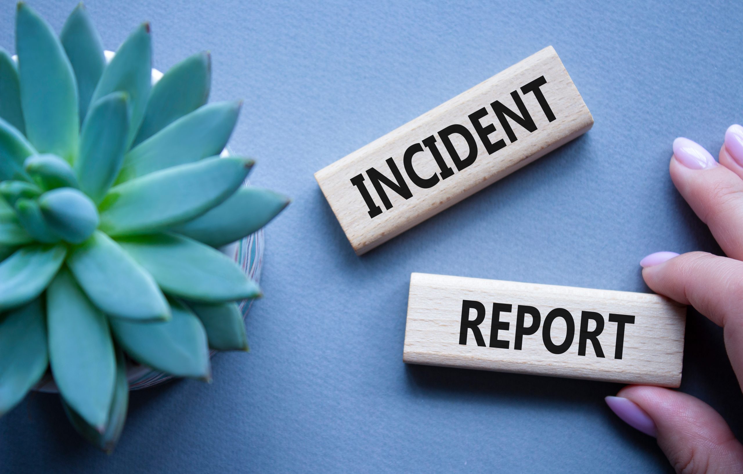 A close-up photo shows two wooden blocks with the words incident reporting written in bold black letters, placed on a light blue background. On the left side, there is a green succulent plant, while on the right, a hand with light purple nail polish is holding one of the blocks.