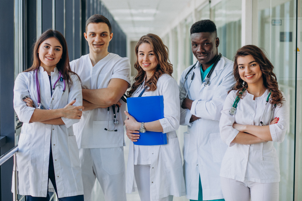 A group of five healthcare professionals stands in a hallway, smiling and posing for the camera. They are wearing white coats and have stethoscopes around their necks, representing different roles in a medical team.
