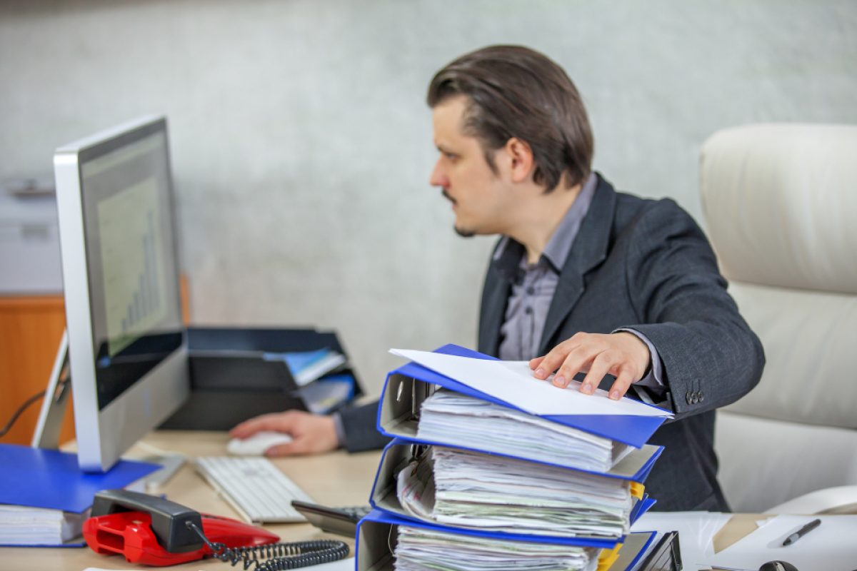 A man in a formal suit is sitting at a desk, focused on a computer screen and thinking “Why is it important to report incidents in the workplace”. The office workspace has various office supplies, including a phone, files, and a computer monitor displaying a graph.