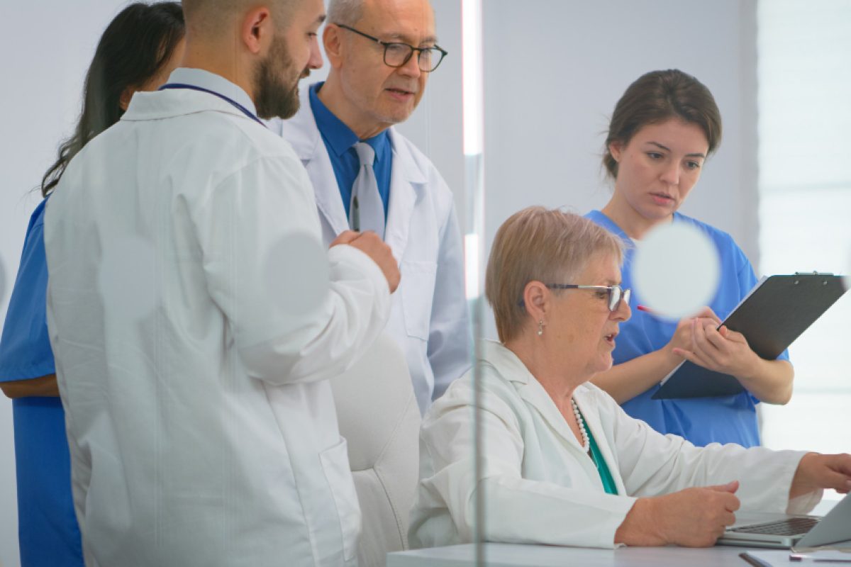 A group of healthcare professionals, including doctors and nurses, gather in a hospital meeting room. One female doctor is seated at a desk, typing on a laptop, while another nurse takes notes on a clipboard. The team appears to be discussing the incident reporting procedure.