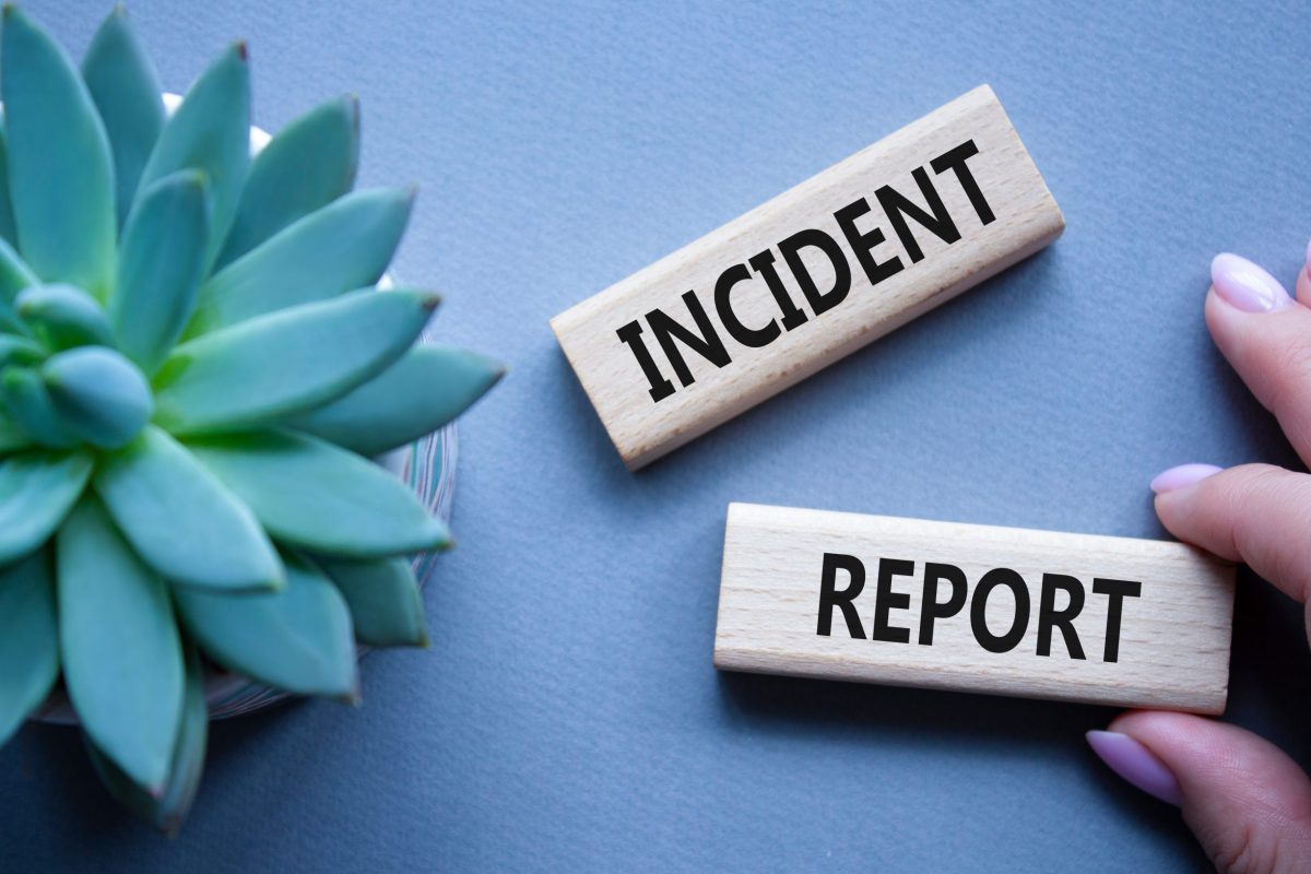 A close-up photo shows two wooden blocks with the words incident reporting written in bold black letters, placed on a light blue background. On the left side, there is a green succulent plant, while on the right, a hand with light purple nail polish is holding one of the blocks.