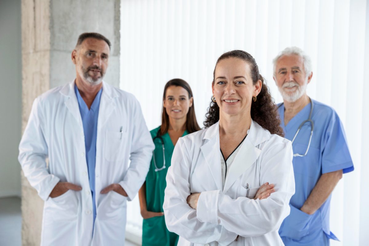 A group of four medical professionals standing together, wearing scrubs and lab coats. The background features large windows with vertical blinds. The team is happy because of the benefits of incident reporting that they had in the hospital.