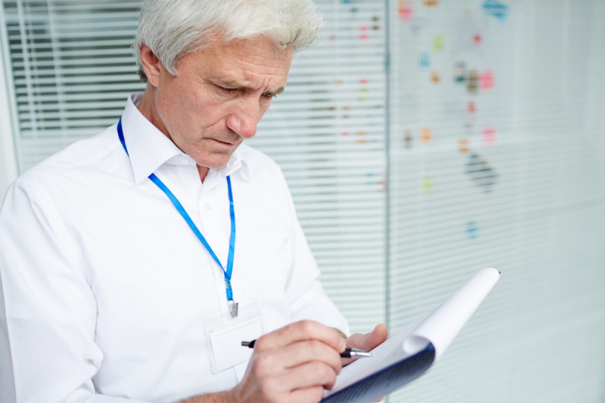 A senior man with gray hair, wearing a white shirt and a blue lanyard, is focused on writing on a notepad. He appears to be reviewing documents related to accreditation compliance in a modern office setting, with glass walls and sticky notes in the background.